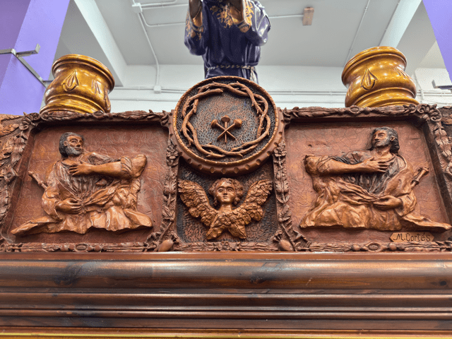 La parte frontal la preside el escudo de la cofradía que consta de los 3 clavos y la corona de espinas, justo debajo un ángel y sus lados dos apóstoles con cetros.